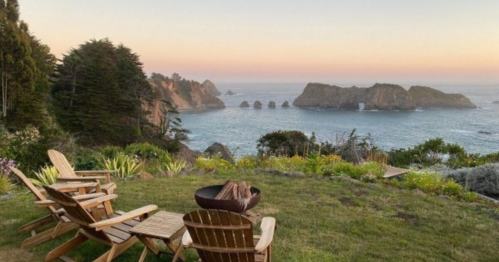 A serene coastal view at sunset, featuring wooden chairs and a fire pit overlooking the ocean and rocky cliffs.