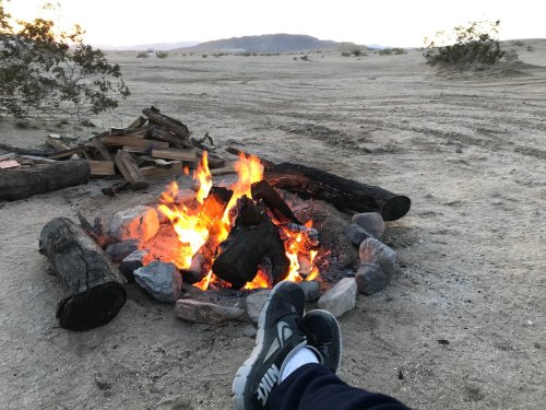 A cozy campfire surrounded by rocks, with a person's feet resting nearby in a desert landscape at dusk.