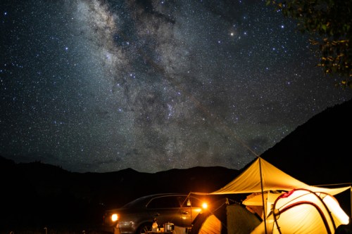 A tent and car under a starry night sky, showcasing the Milky Way and distant mountains.