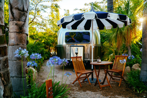 A cozy outdoor scene featuring an Airstream trailer, a table with chairs, and lush greenery with blue flowers.