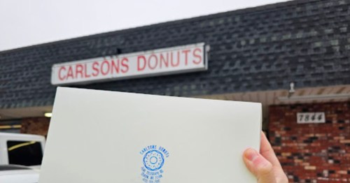 A hand holds a white box with a blue logo in front of Carlson's Donuts sign.
