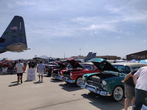 A row of classic cars with hoods open at an outdoor event, with people walking and military aircraft in the background.