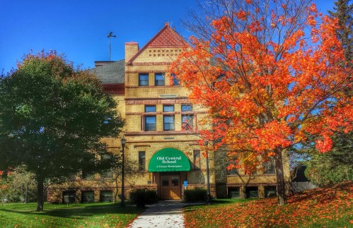 Historic brick school building with a green sign, surrounded by vibrant autumn trees and a clear blue sky.