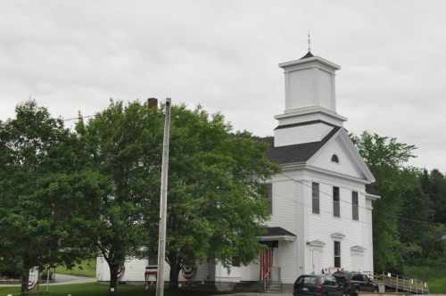 A white church building with a tall steeple, surrounded by green trees and a cloudy sky.