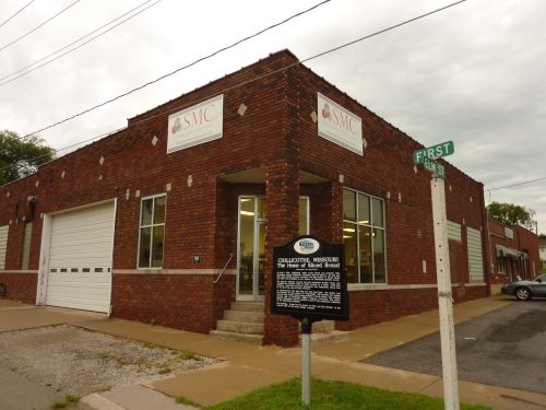 Brick building with signs for "SMC" and a historical marker at the corner of First Street. Overcast sky above.