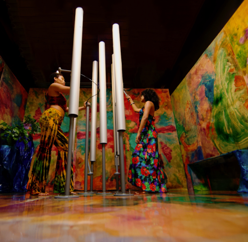 Two women in colorful outfits interact with tall, white musical tubes in a vibrant, painted room.