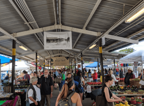 A bustling market scene with people browsing stalls under a covered area, colorful tents, and fresh produce on display.