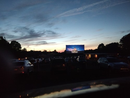 A drive-in movie theater at dusk, with cars parked and a large screen displaying a film against a colorful sky.