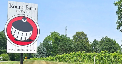 Sign for Round Barn Estate with a round barn logo, surrounded by green vineyards and trees on a sunny day.