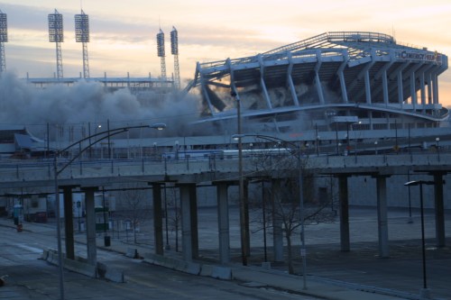 Demolition of a stadium, with smoke rising and structures collapsing under a cloudy sky at dawn.
