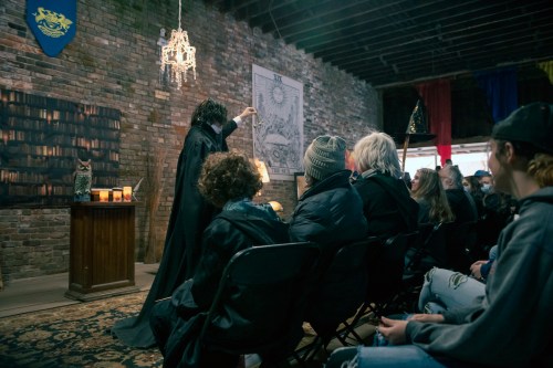 A performer in a cloak holds an object, captivating an audience seated in a rustic, brick-walled venue.