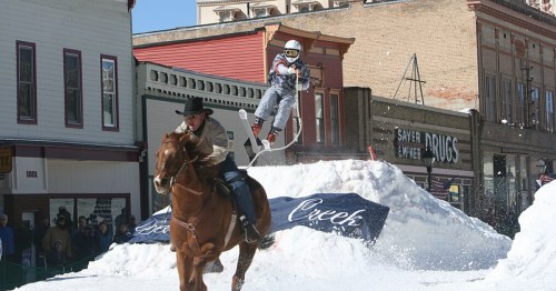 A skier jumps off a snow ramp while a cowboy rides a horse nearby, with buildings in the background.