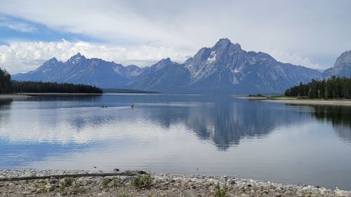 A serene lake reflects majestic mountains under a partly cloudy sky, surrounded by lush greenery.