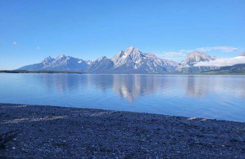 A serene lake reflects majestic mountains under a clear blue sky, with a pebbly shore in the foreground.