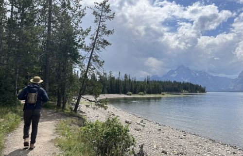 A person walks along a lakeside trail, surrounded by trees and mountains under a partly cloudy sky.