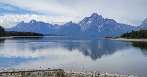 A serene lake reflects majestic mountains under a partly cloudy sky, surrounded by lush greenery.