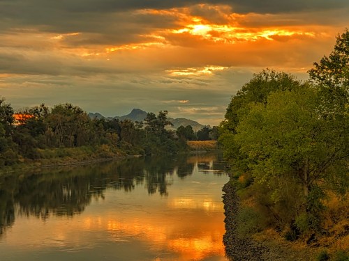 A serene river reflects a vibrant sunset, surrounded by trees and distant mountains under a cloudy sky.