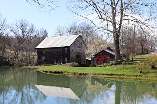 A rustic wooden building by a calm pond, reflecting trees and a blue sky, with a small house nearby.