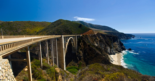 A scenic view of a coastal bridge spanning a cliffside, with mountains and the ocean in the background under a clear blue sky.