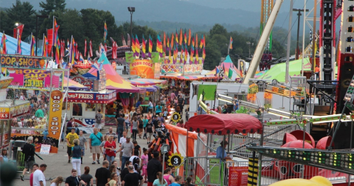 A vibrant fair scene with colorful flags, food stalls, and crowds enjoying rides and games under a clear sky.
