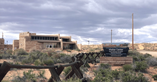 Visitor center building with stone architecture, surrounded by desert landscape and cloudy skies. Wooden fence in foreground.