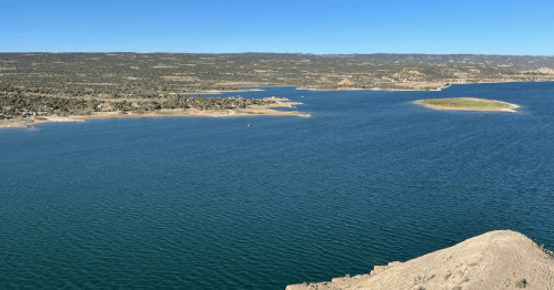 A serene view of a blue lake surrounded by hills and a small island under a clear blue sky.