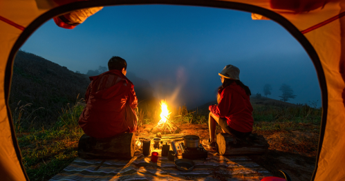 Two campers sit by a campfire outside their tent, surrounded by nature at dusk.