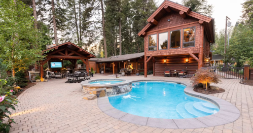 A cozy wooden house with a pool and hot tub, surrounded by trees and outdoor seating areas.