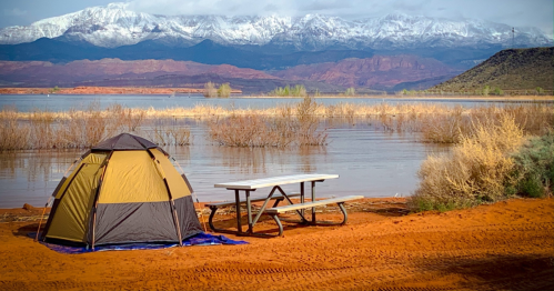 A tent and picnic table by a lake, with snow-capped mountains in the background and orange sandy terrain.