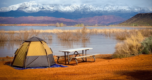 A tent and picnic table by a lake, with snow-capped mountains in the background and orange sandy terrain.