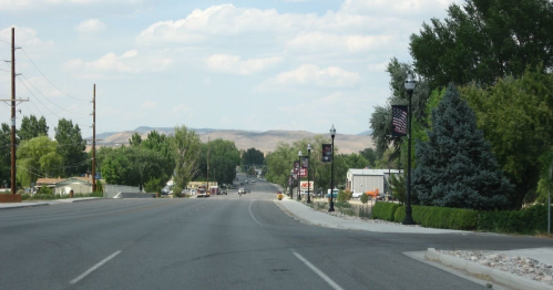 A quiet road lined with trees and banners, leading towards distant hills under a partly cloudy sky.