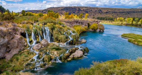 A scenic view of a waterfall cascading into a river, surrounded by lush greenery and rocky terrain under a blue sky.