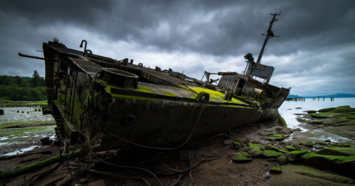 A weathered shipwreck covered in moss, leaning on a rocky shore under a cloudy sky.