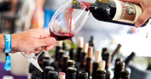 A hand pours red wine from a bottle into a glass at a wine tasting event, surrounded by various wine bottles.