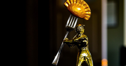 A golden figurine holding a fork with a golden empanada balanced on top, set against a blurred background.