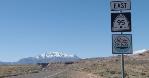 A road sign for East 95 with a scenic byway logo, set against a backdrop of snow-capped mountains and clear blue sky.