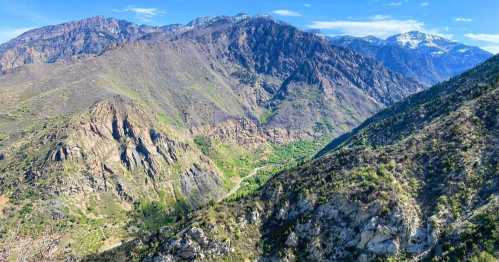 A panoramic view of rugged mountains with green valleys and snow-capped peaks under a clear blue sky.