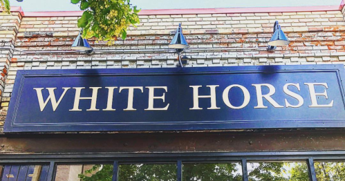 Sign for "White Horse" displayed on a brick wall with outdoor lighting fixtures above it.
