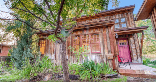 A rustic wooden house surrounded by greenery and trees, featuring large windows and a red door.