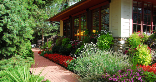 A vibrant garden with colorful flowers and a brick pathway leading to a charming house surrounded by trees.