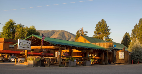 A rustic wooden building with a green roof, surrounded by trees and mountains, featuring outdoor seating and American flags.