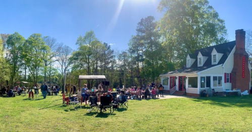 A sunny outdoor gathering with people socializing near a house, surrounded by trees and chairs set up on the lawn.