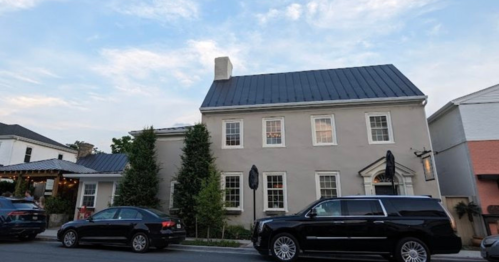 A two-story house with a gray exterior and a metal roof, surrounded by trees and parked cars on a street.