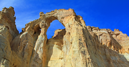 A natural rock arch stands tall against a bright blue sky, surrounded by rugged sandstone cliffs.