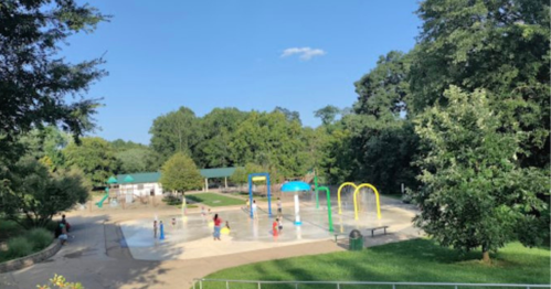 A sunny park with a splash pad, colorful play structures, and children playing among trees and greenery.