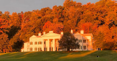 A grand white mansion surrounded by vibrant autumn foliage and a grassy lawn, illuminated by warm sunset light.
