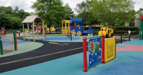 Colorful playground with various play structures, including a train and a yellow truck, surrounded by green trees.