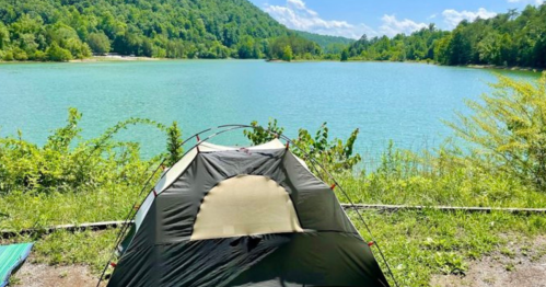 A tent set up by a serene lake, surrounded by lush greenery and mountains under a clear blue sky.
