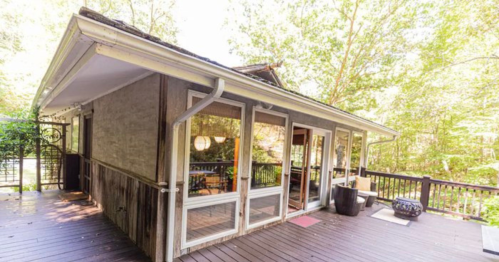 A cozy cabin with large windows, surrounded by trees, featuring a wooden deck and outdoor seating area.