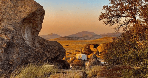 A scenic landscape featuring large rocks, distant mountains, and an RV surrounded by grass at sunset.